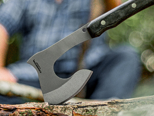 Condor hatchet bitten into a fallen log in the forest, with a man in a plaid shirt sitting in the background
